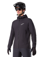 Alpinestars A-Dura Thermal Jacket, giacca da ciclismo nera, design con cappuccio e zip frontale, logo a stella bianco sul petto, indossata con casco da mountain bike e guanti neri, abbigliamento sportivo per il trail riding in climi freddi