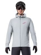 Alpinestars A-Dura Thermal Jacket in Titanium, giacca da ciclismo grigio chiaro con cappuccio e logo a stella rosso sul petto, indossata da un rider con casco da MTB nero, occhiali da sole e guanti, abbigliamento isolante per mountain bike