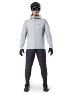 Giacca Termica Alpinestars A-Dura, grigio titanio, giacca da ciclismo indossata con pantaloni da mountain bike neri, guanti e casco, caratterizzata da un piccolo logo a stella rosso sul petto e cerniera frontale completa, abbigliamento isolante leggero per la guida fuoristrada