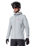 Alpinestars A-Dura Thermal Jacket, giacca termica da ciclismo grigio chiaro, design con cappuccio e piccolo logo rosso, indossata con casco e guanti neri, abbigliamento tecnico da mountain bike per il freddo