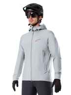 Alpinestars A-Dura Thermal Jacket, giacca termica da ciclismo grigio chiaro, design con cappuccio e piccolo logo rosso, indossata con casco e guanti neri, abbigliamento tecnico da mountain bike per il freddo