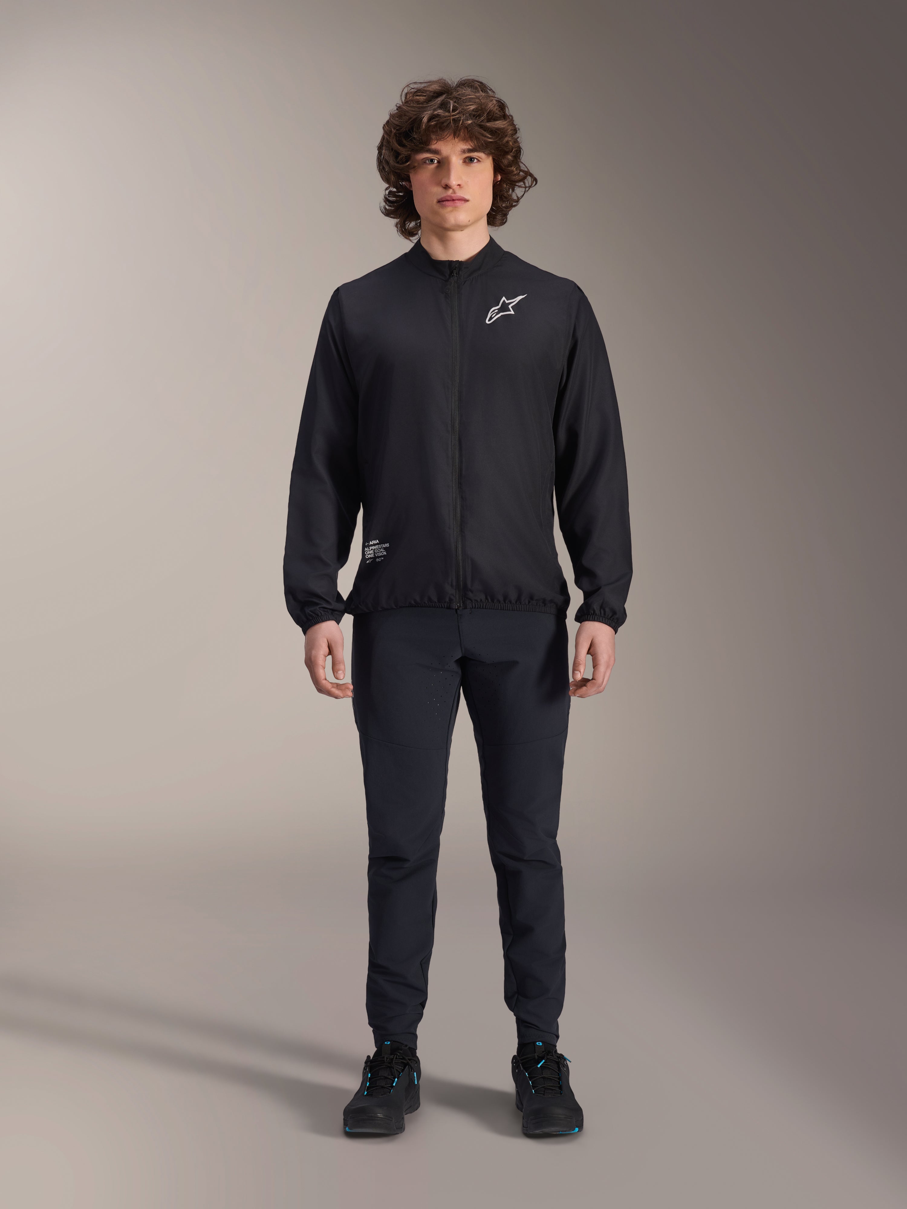 Modello che indossa la giacca frangivento Alpinestars A-Aria in nero, con cerniera frontale integrale, collo alto e logo Astars bianco sul petto, abbinata a pantaloni tecnici neri e scarpe nere, abbigliamento tecnico da ciclismo per mountain bike e uso outdoor.