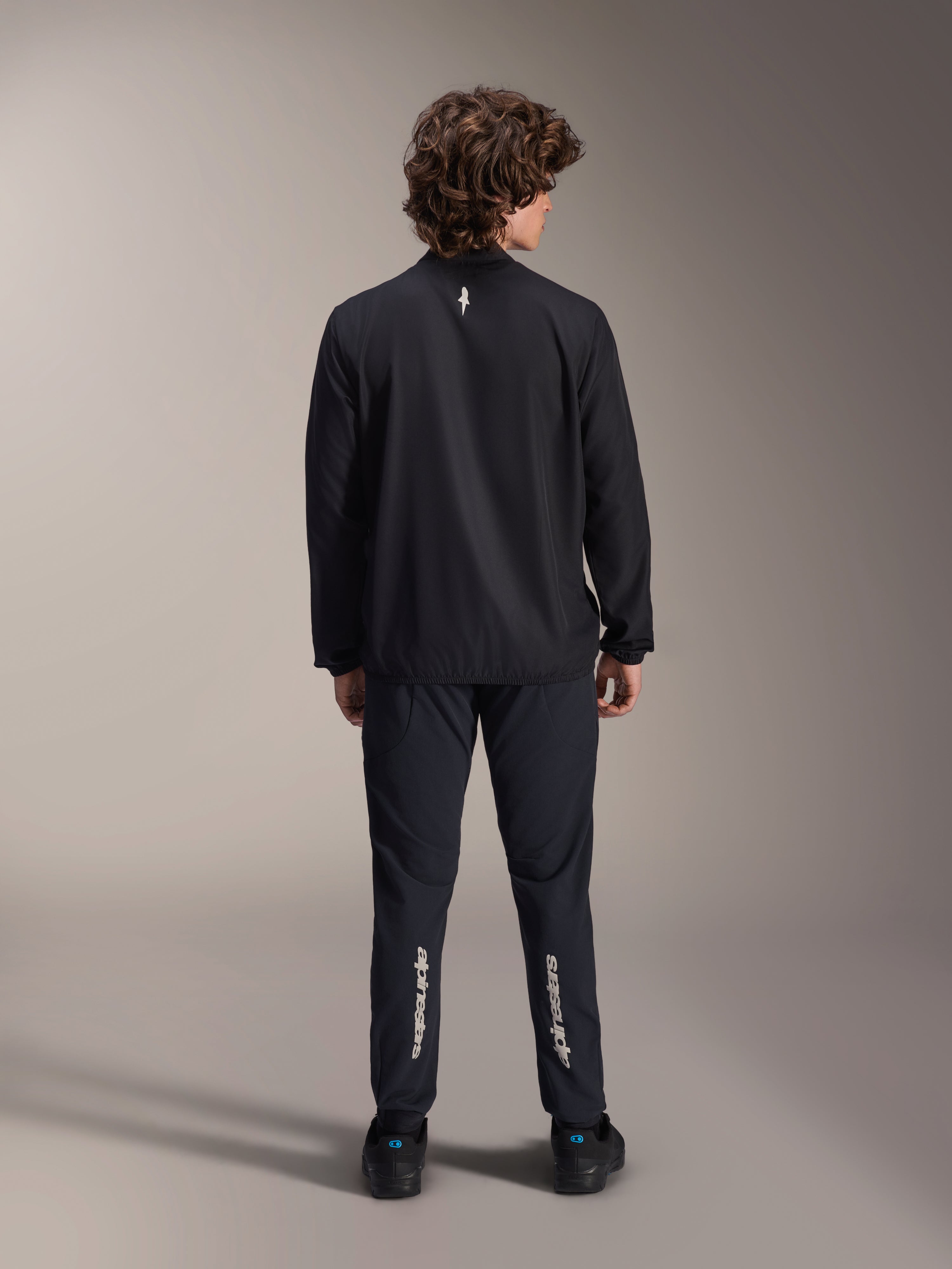 Giacca frangivento Alpinestars A-Aria, nero, vista posteriore, giacca da ciclismo leggera e resistente al vento con un piccolo logo bianco sulla parte superiore della schiena, indossata con pantaloni neri coordinati con marchio Alpinestars verticale sui polpacci, abbigliamento tecnico da mountain bike