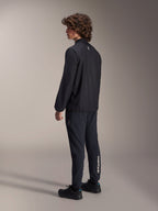 Giacca frangivento Alpinestars A-Aria, nera, giacca da ciclismo leggera e resistente al vento, vista posteriore di tre quarti su modello, con un piccolo logo bianco sul retro del collo e bordo elasticizzato, abbinata a pantaloni e scarpe neri coordinati per un look orientato alle prestazioni.