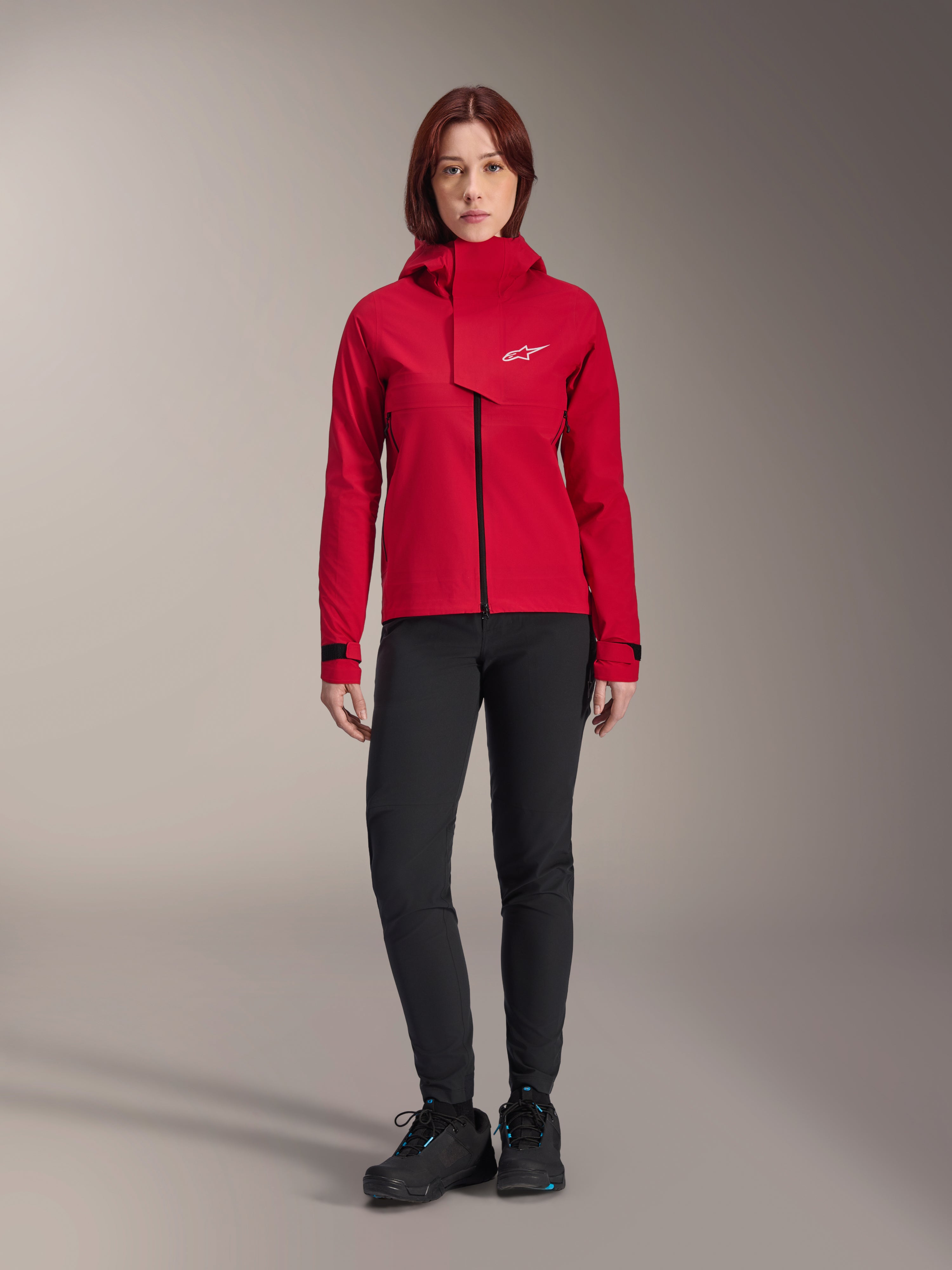 Giacca impermeabile Alpinestars Stella A-Dura Elite, giacca da MTB per donna, Rosso Fluo, indossata con pantaloni e scarpe neri, con cappuccio a collo alto e logo Alpinestars bianco, abbigliamento tecnico da mountain bike