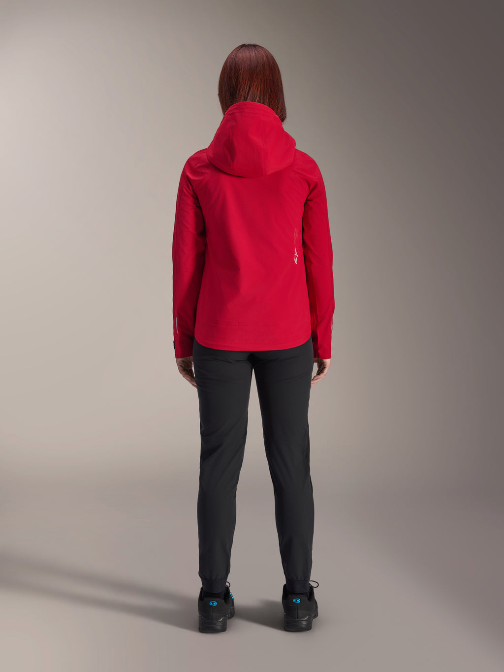 Vista posteriore di una donna che indossa la giacca impermeabile Alpinestars Stella A-Dura Elite in Red Fluo, abbinata a pantaloni e scarpe neri, dotata di un cappuccio protettivo e orlo posteriore allungato per la mountain bike.