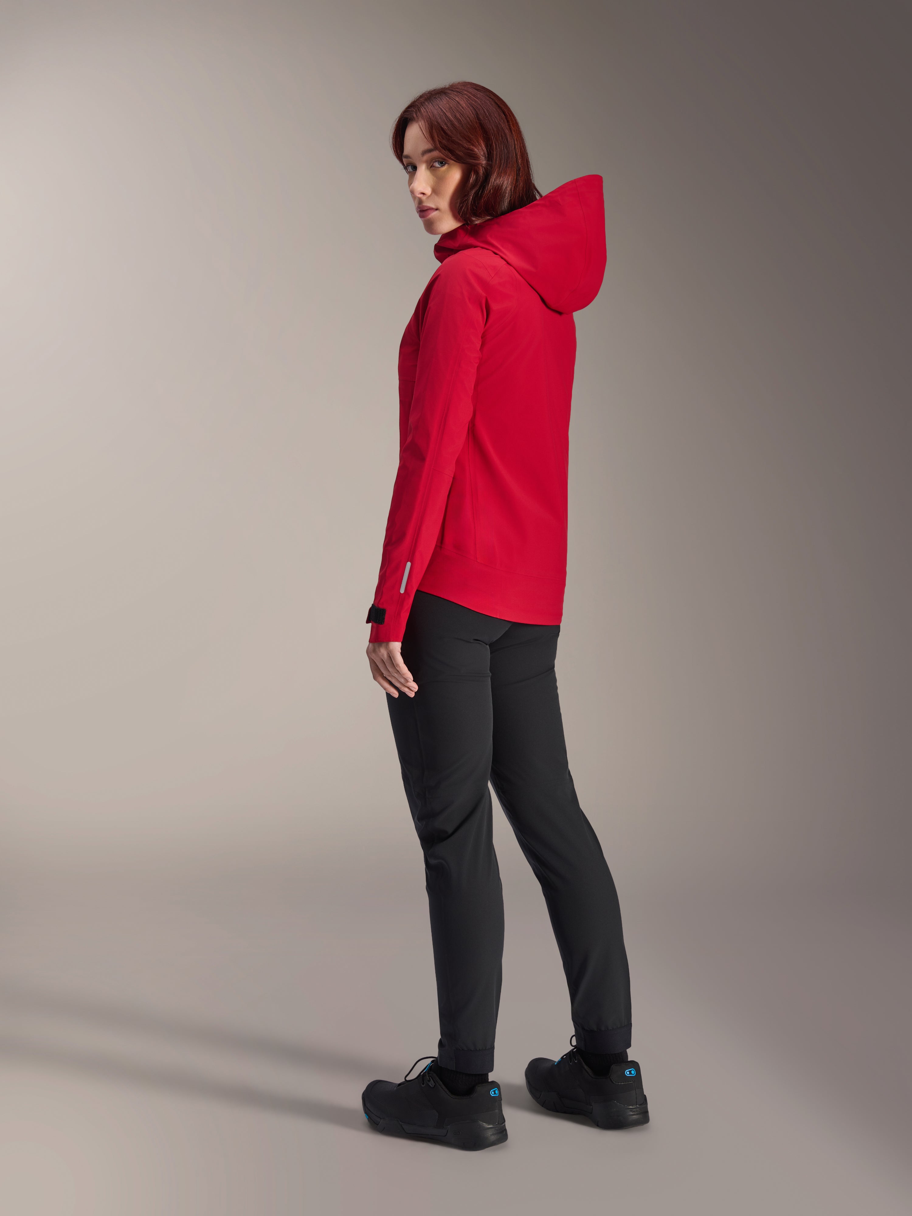 Donna che indossa la giacca impermeabile Alpinestars Stella A-Dura Elite in Red Fluo, abbinata a pantaloni tecnici da riding neri e scarpe nere, vista posteriore di tre quarti che mostra il cappuccio regolabile e l'orlo posteriore allungato, abbigliamento tecnico per mountain bike