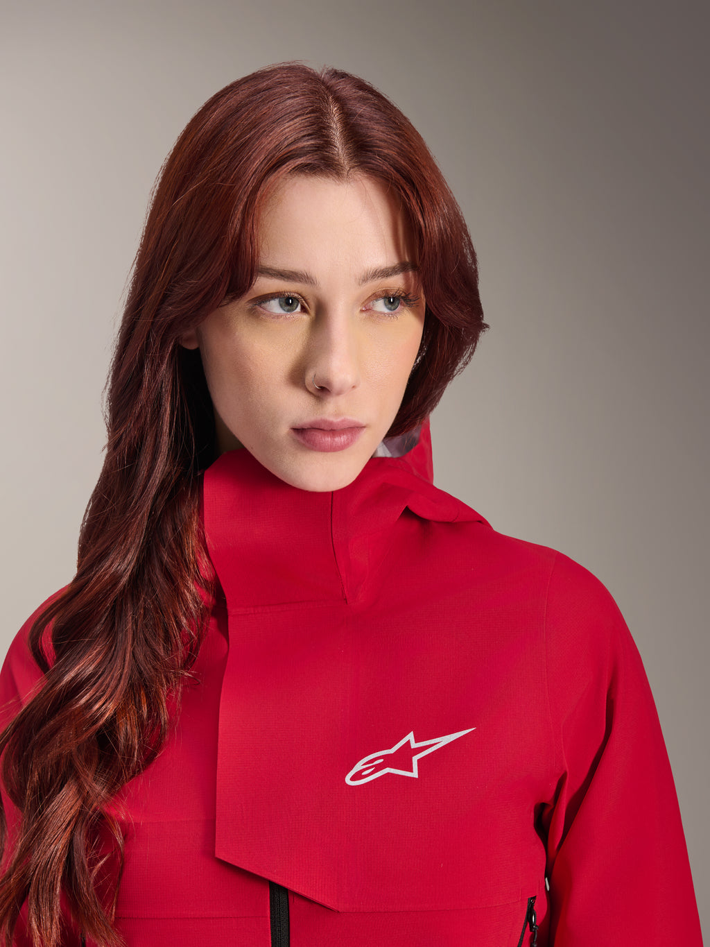 Alpinestars Stella A-Dura Elite Waterproof Jacket, rosso fluo, indossata da una donna con lunghi capelli rossi, giacca tecnica impermeabile a 3 strati con collo alto e logo bianco, equipaggiamento tecnico per outdoor e mountain bike