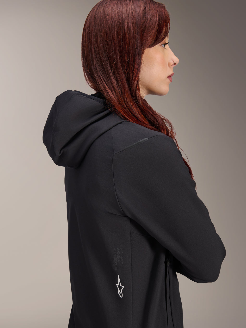 Giacca Termica Alpinestars Stella A-Dura, giacca da ciclismo da donna, nero, profilo laterale di una donna con i capelli rossi che indossa la giacca, dotata di cappuccio integrato e logo a stella Alpinestars bianco sul lato, abbigliamento termico isolante leggero per MTB