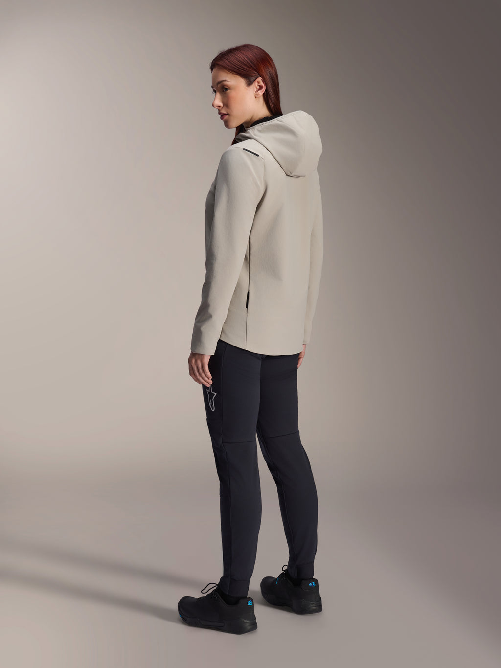 Giacca termica Alpinestars Stella A-Dura in grigio titanio, vista posteriore a tre quarti di una donna che indossa la giacca da ciclismo con cappuccio e pantaloni da MTB neri, scatto in studio con dettaglio della tasca