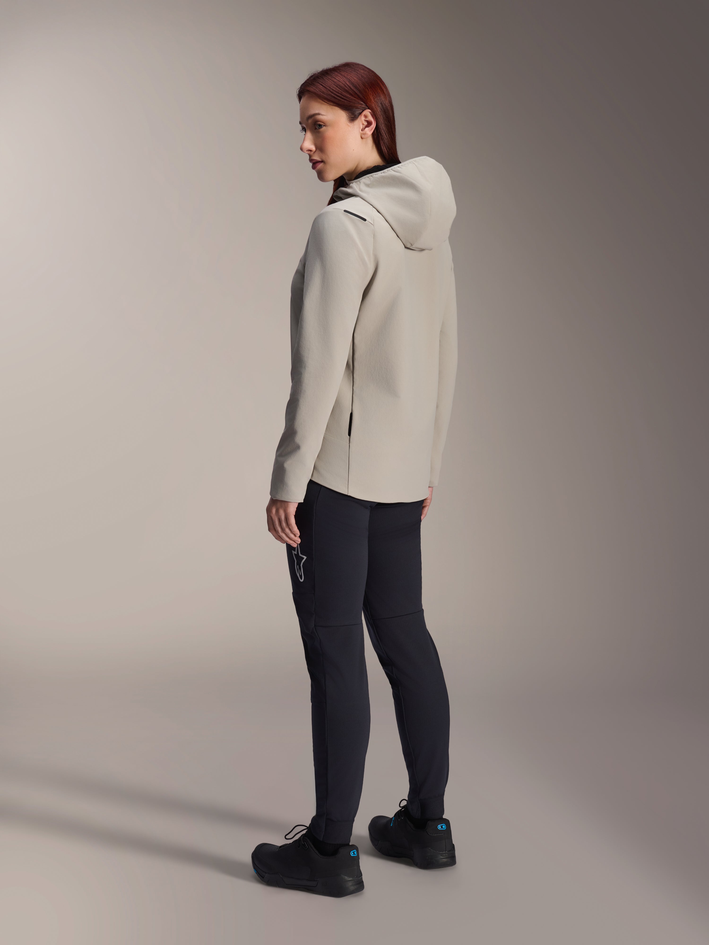 Giacca termica Alpinestars Stella A-Dura in grigio titanio, vista posteriore a tre quarti di una donna che indossa la giacca da ciclismo con cappuccio e pantaloni da MTB neri, scatto in studio con dettaglio della tasca