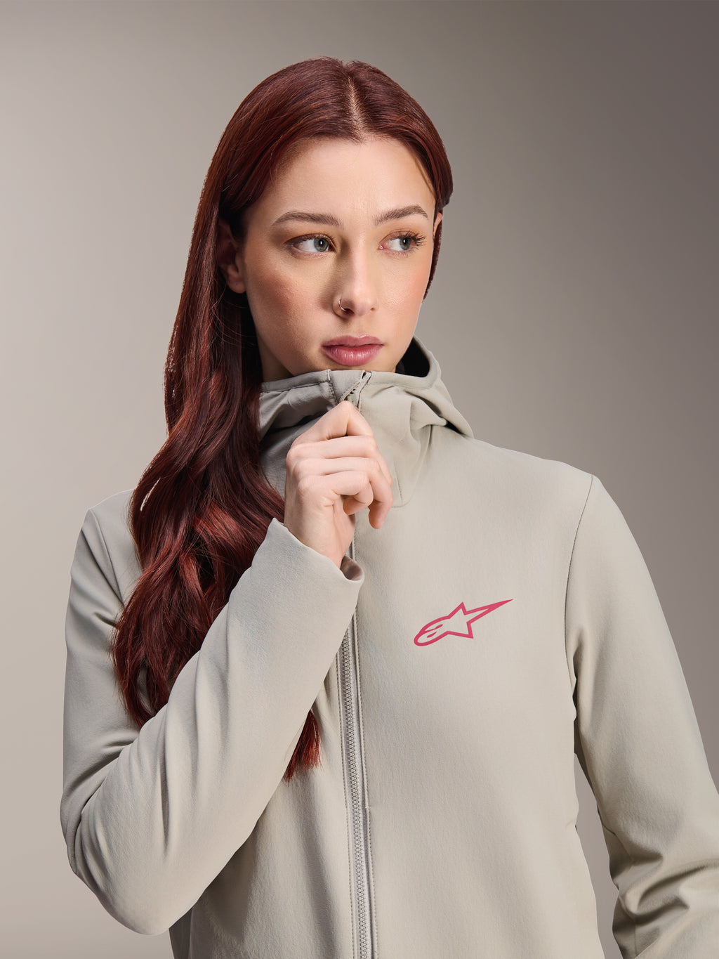 Giacca termica da donna Alpinestars Stella A-Dura, grigio titanio con logo Alpinestars a stella rosa, modella con lunghi capelli rossi che indossa e chiude la zip della giacca con cappuccio, abbigliamento da ciclismo leggero e isolante per il freddo