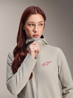 Giacca termica da donna Alpinestars Stella A-Dura, grigio titanio con logo Alpinestars a stella rosa, modella con lunghi capelli rossi che indossa e chiude la zip della giacca con cappuccio, abbigliamento da ciclismo leggero e isolante per il freddo