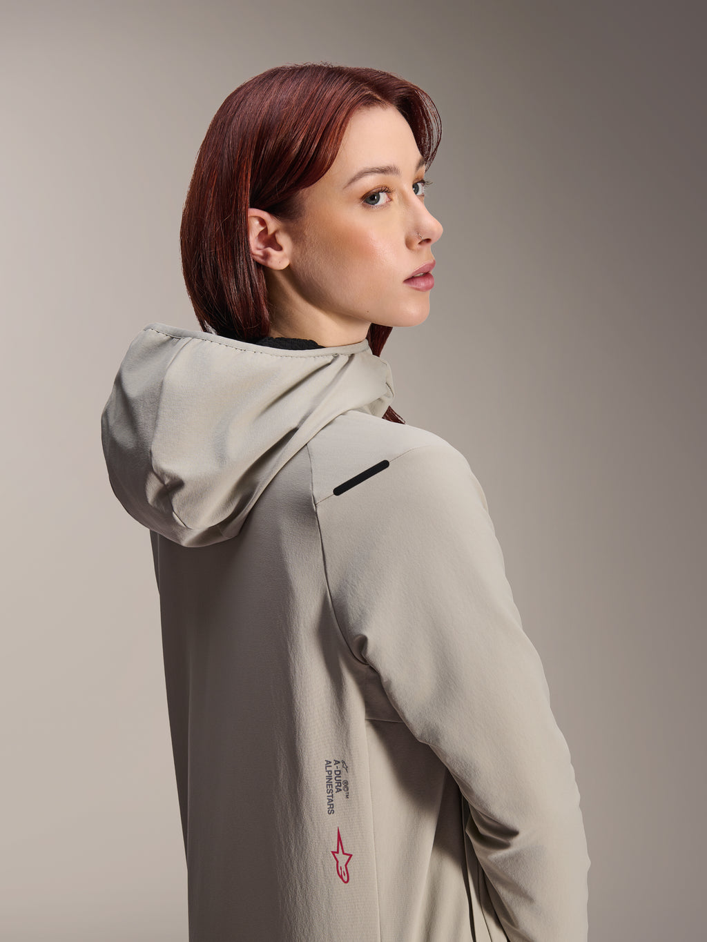 Alpinestars Stella A-Dura Thermal Jacket, giacca da MTB da donna, grigio titanio, donna che guarda oltre la spalla indossando una giacca tecnica con cappuccio, con scritta Alpinestars e logo a stella rosso sul lato, progettata per il ciclismo in climi freddi