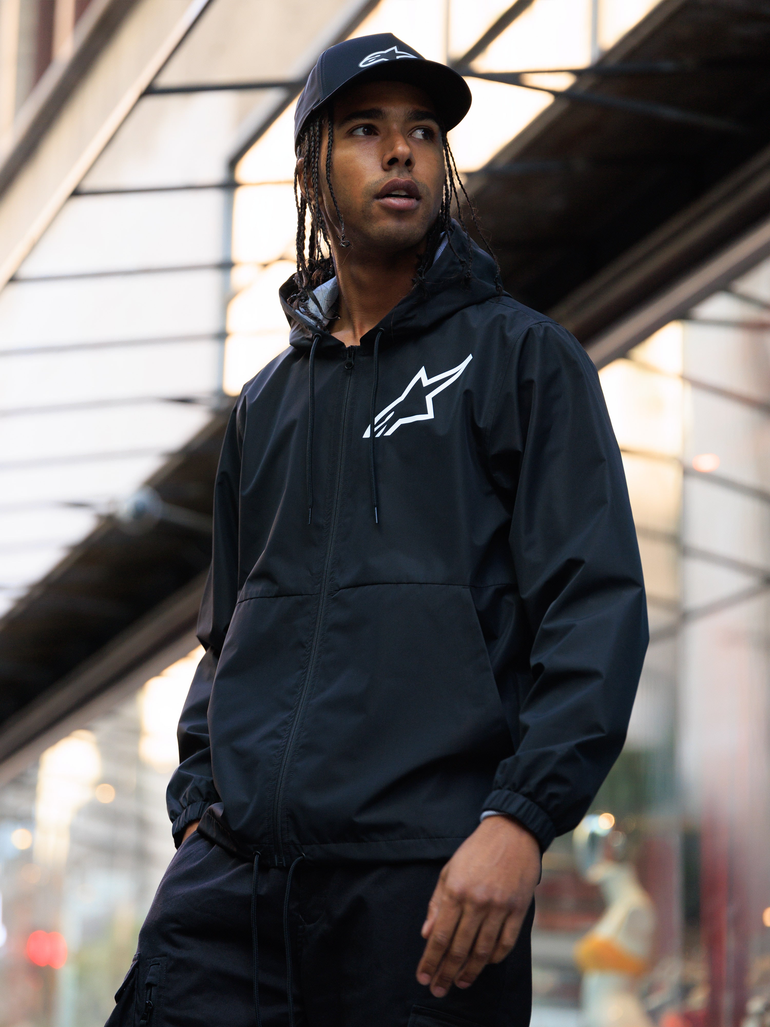 Giacca a vento Alpinestars Speeder Chromium, nera, giacca a vento con cappuccio e logo bianco sul petto, indossata da un uomo con cappellino Alpinestars nero coordinato in un contesto urbano, abbigliamento casual motorsport streetwear.