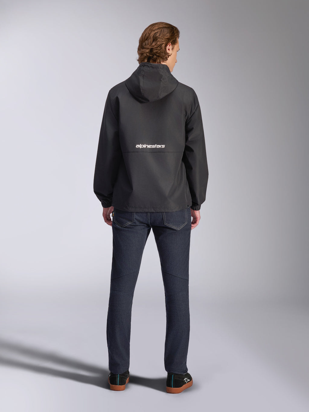 Giacca frangivento Alpinestars Speeder Chromium, nero, vista posteriore con marchio Alpinestars bianco al centro, modello con cappuccio, indossata da un modello con pantaloni in denim scuro, giacca casual ispirata al motorsport