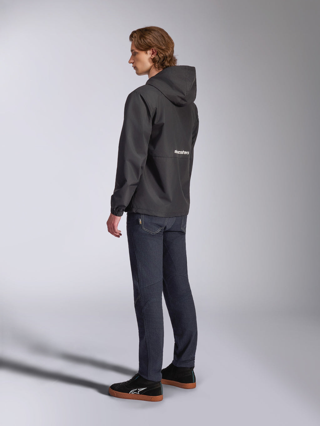 Giacca a vento Alpinestars Speeder Chromium, nera con logo bianco sul retro, giacca casual da uomo con cappuccio, vista di profilo indossata con jeans scuri e sneakers nere, streetwear ispirato al motorsport