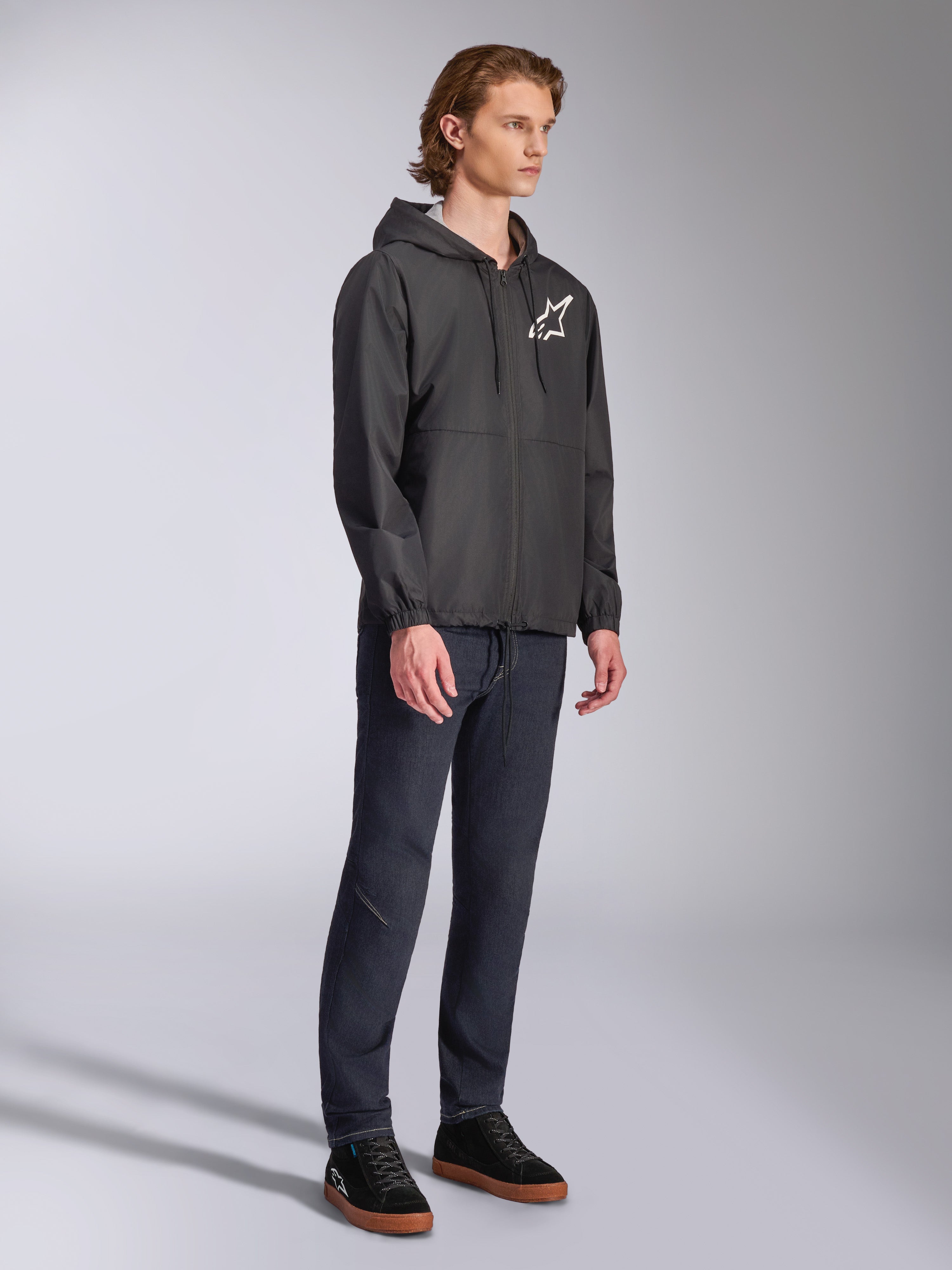 Alpinestars Speeder Chromium Windbreak Jacket, giacca frangivento nera con cappuccio e cerniera anteriore, logo a stella Alpinestars bianco sul lato sinistro del petto, streetwear casual motorsport, abbinata a pantaloni scuri e sneakers nere in studio.