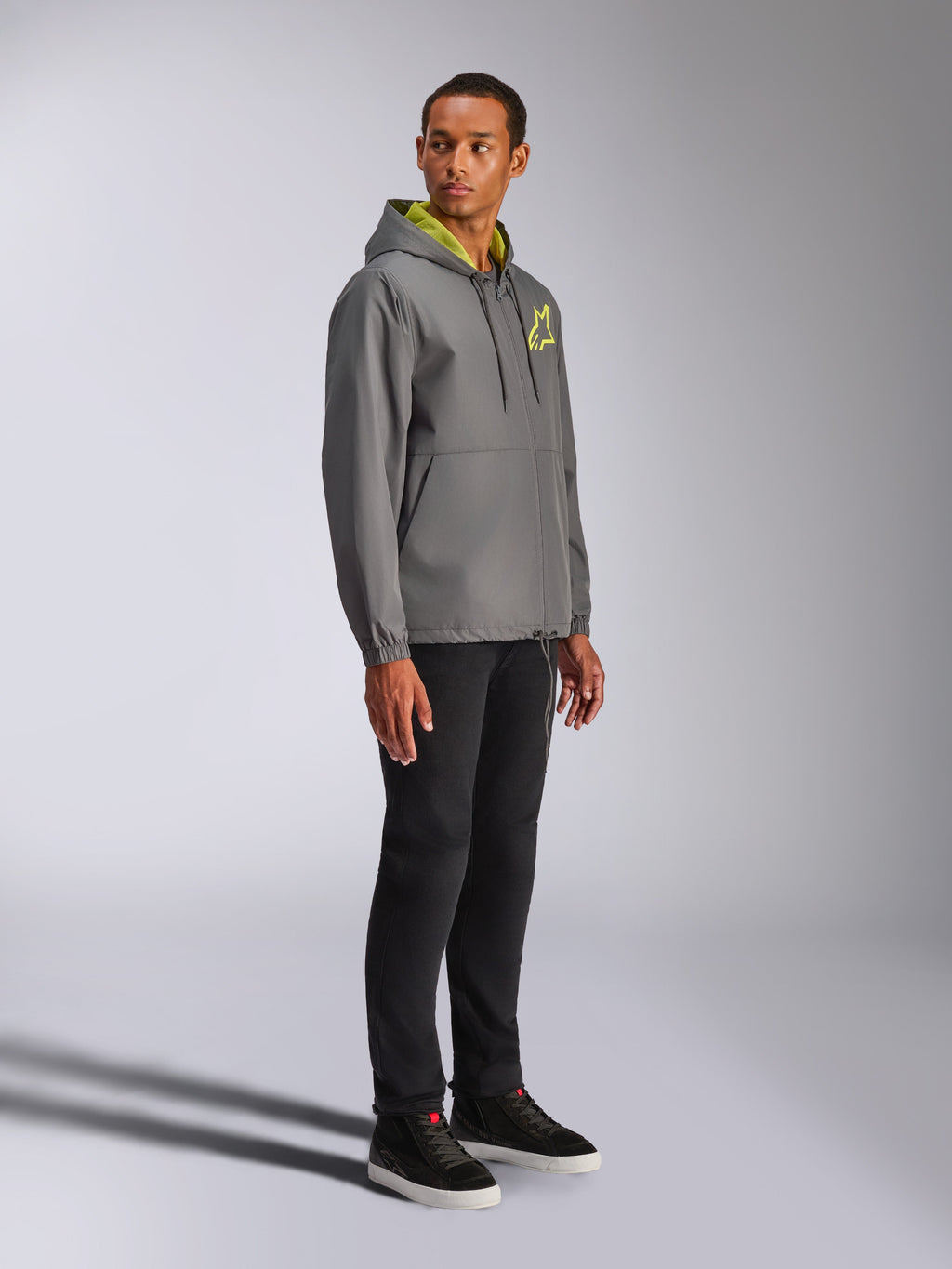 Giacca frangivento Alpinestars Speeder Chromium, antracite con dettagli giallo fluo, modello che indossa la giacca con cappuccio, logo a stella giallo sul petto, abbinata a pantaloni e sneakers nere, streetwear casual ispirato al motorsport
