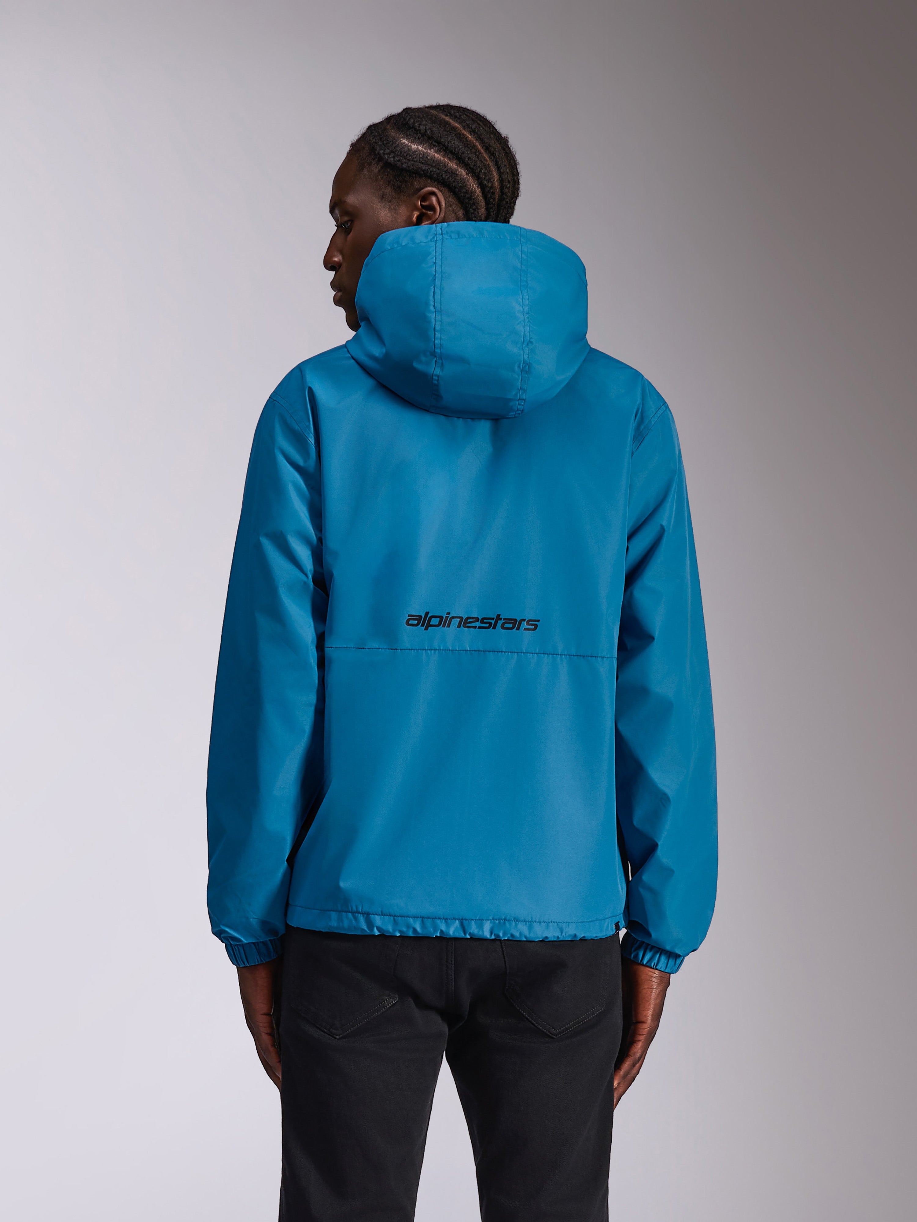 Giacca a vento Alpinestars Speeder Chromium, blu, vista posteriore su modello con logo a scritta nero sul retro, giacca leggera con cappuccio e polsini elastici, abbigliamento sportivo casual.