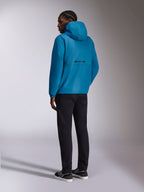 Giacca a vento Alpinestars Speeder Chromium, giacca a vento con cappuccio, blu, vista posteriore con logo Alpinestars nero sul retro, indossata con pantaloni neri e sneakers bianche e nere, streetwear casual ispirato al motorsport