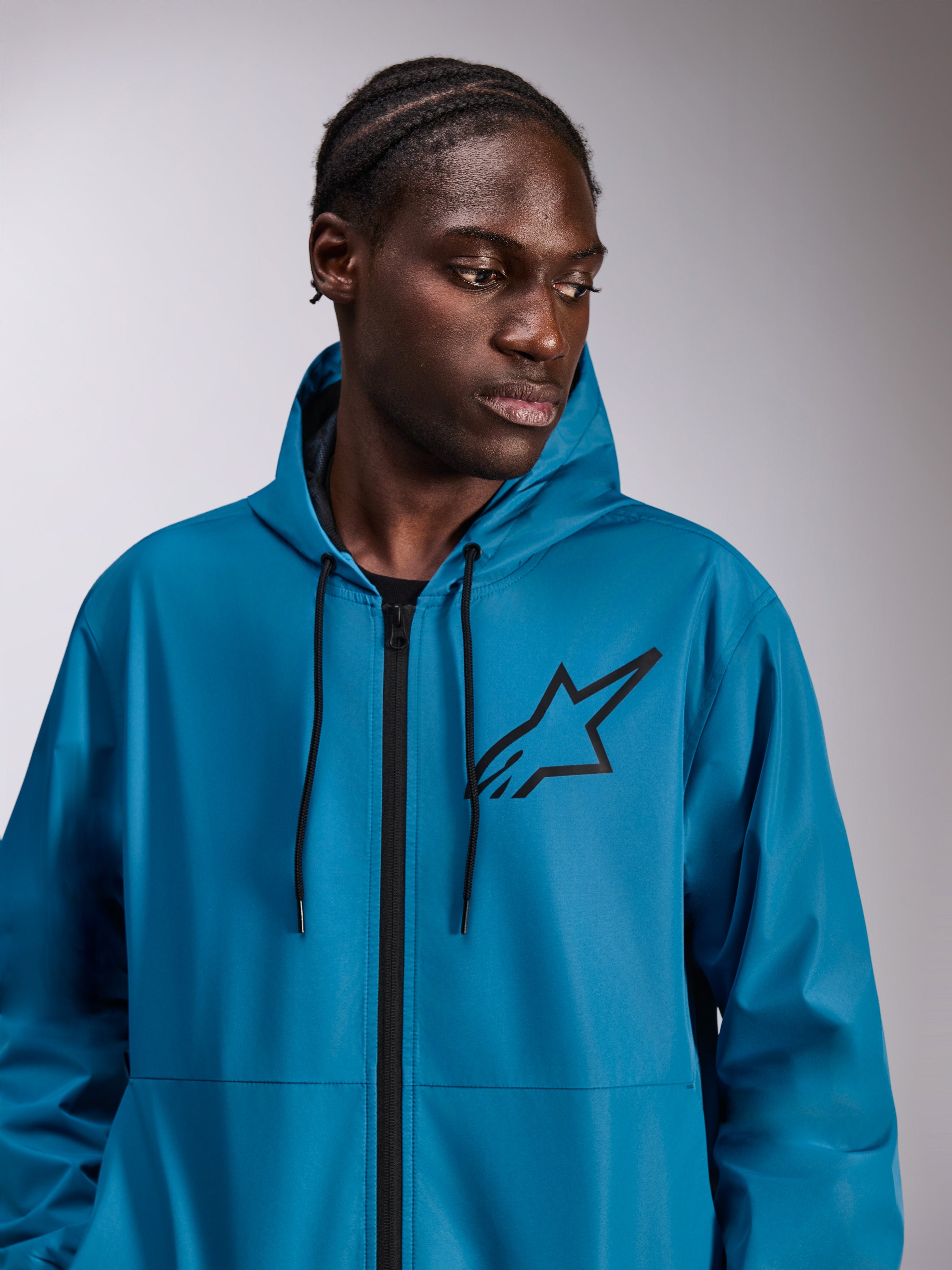 Modello che indossa la giacca a vento Alpinestars Speeder Chromium, blu, giacca a vento leggera con cappuccio, logo Astars nero sul petto, cerniera anteriore e cordini neri, streetwear casual ispirato al motorsport