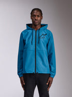 Alpinestars Speeder Chromium Windbreak Jacket, giacca a vento blu con cappuccio e logo Astars nero sul petto, cerniera frontale a tutta lunghezza, polsini elastici e cappuccio con coulisse, indossata da un modello come abbigliamento sportivo casual.