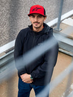 Uomo che indossa la giacca Alpinestars Overhaul Racer in nero, una giacca guscio antipioggia con cappuccio resistente all'acqua e piccolo logo sul petto, abbinata a un cappellino Alpinestars rosso, abbigliamento lifestyle casual motorsport