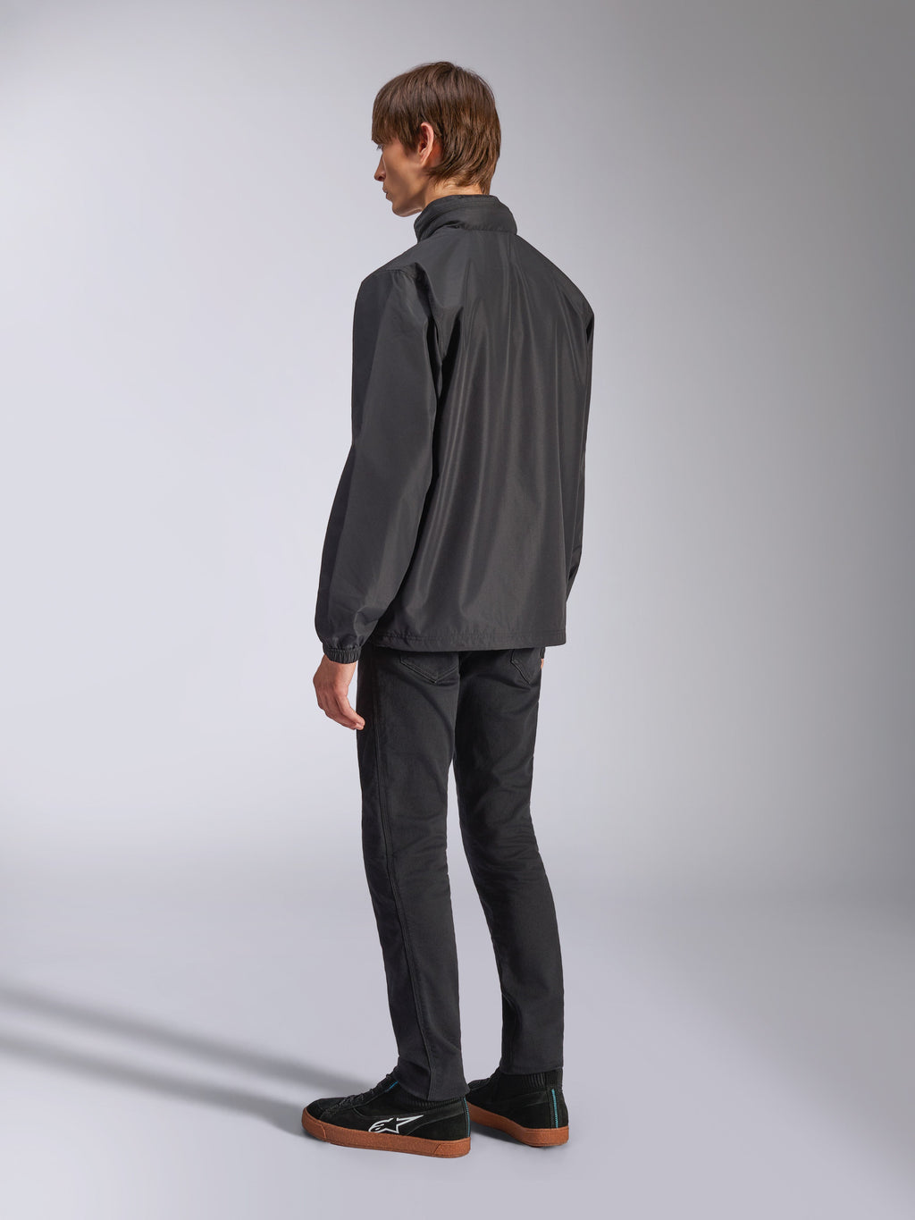 Alpinestars Overhaul Racer Jacket, nera, vista posteriore a tre quarti, indossata da un modello con jeans neri e sneakers nere, giacca guscio antipioggia leggera e resistente all'acqua per uno stile di vita motorsport casual