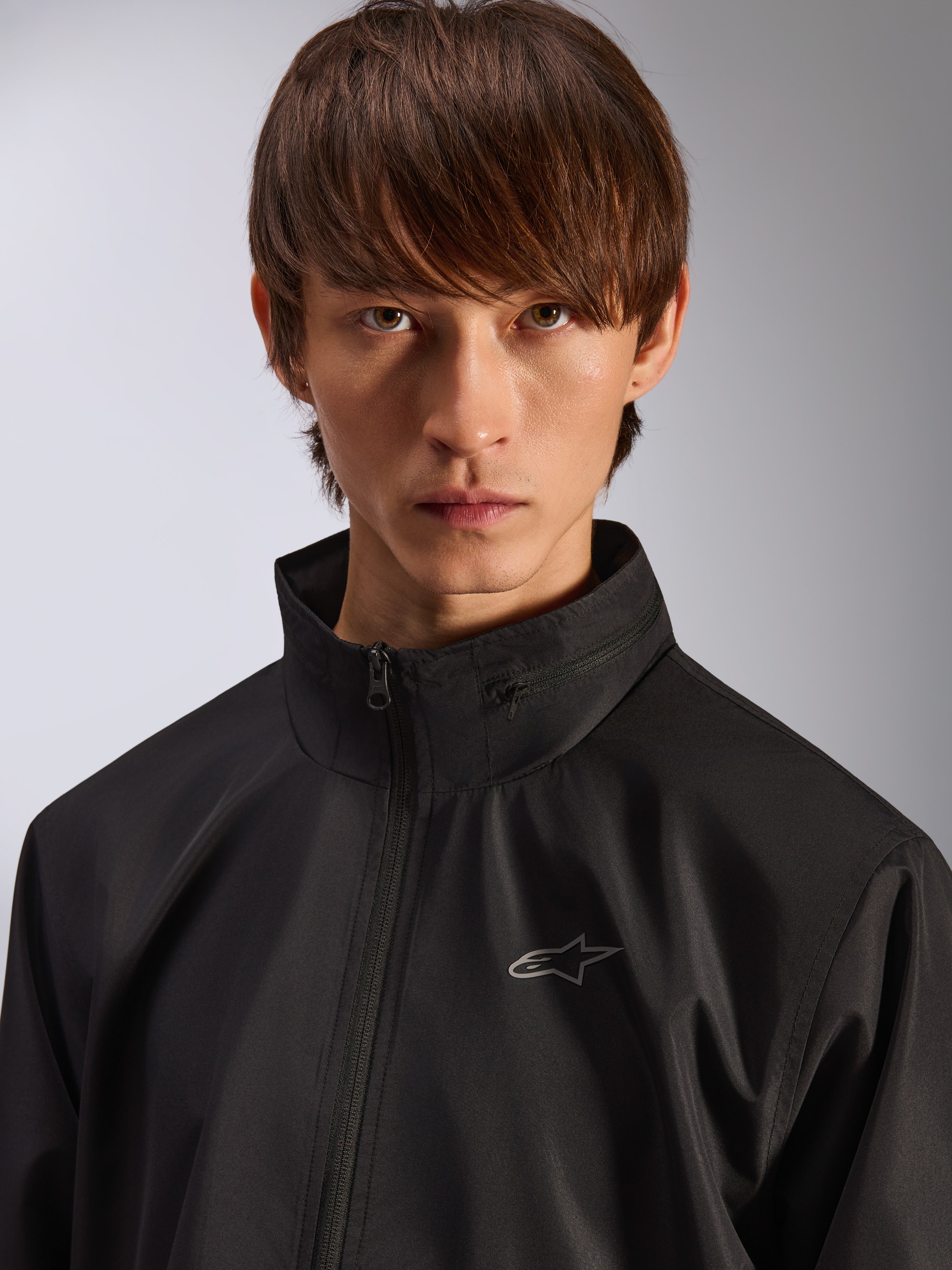 Alpinestars Overhaul Racer Jacket, giacca guscio antipioggia nera, ritratto ravvicinato di un modello che indossa la giacca, con colletto alto e piccolo logo Astars argento sul petto, abbigliamento casual ispirato al motorsport
