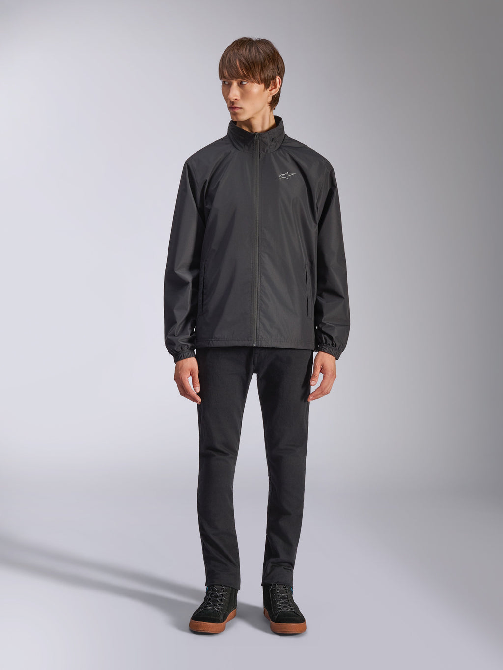 Giacca Alpinestars Overhaul Racer, guscio antipioggia nero da uomo, vista a figura intera su un modello abbinata a pantaloni neri e sneakers casual, con un piccolo logo bianco sul petto, tessuto resistente all'acqua e design casual ispirato al motorsport
