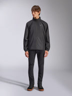 Giacca Alpinestars Overhaul Racer, guscio antipioggia nero da uomo, vista a figura intera su un modello abbinata a pantaloni neri e sneakers casual, con un piccolo logo bianco sul petto, tessuto resistente all'acqua e design casual ispirato al motorsport