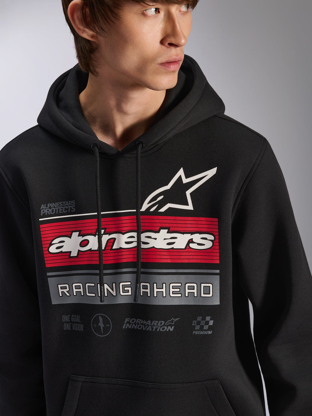 Alpinestars Harken Hoodie, felpa nera con cappuccio in pile indossata da un modello, caratterizzata da una grande grafica sul petto con strisce rosse e grigie, marchio Alpinestars bianco e logo a stella, scritta 'Racing Ahead', streetwear casual moderno ispirato al motorsport.