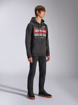 Alpinestars Harken Hoodie, felpa in pile nera con cappuccio, con una grande grafica sul petto con scritta Alpinestars rossa e logo a stella, tasca a marsupio, indossata da un modello con pantaloni neri e sneakers, streetwear casual motorsport