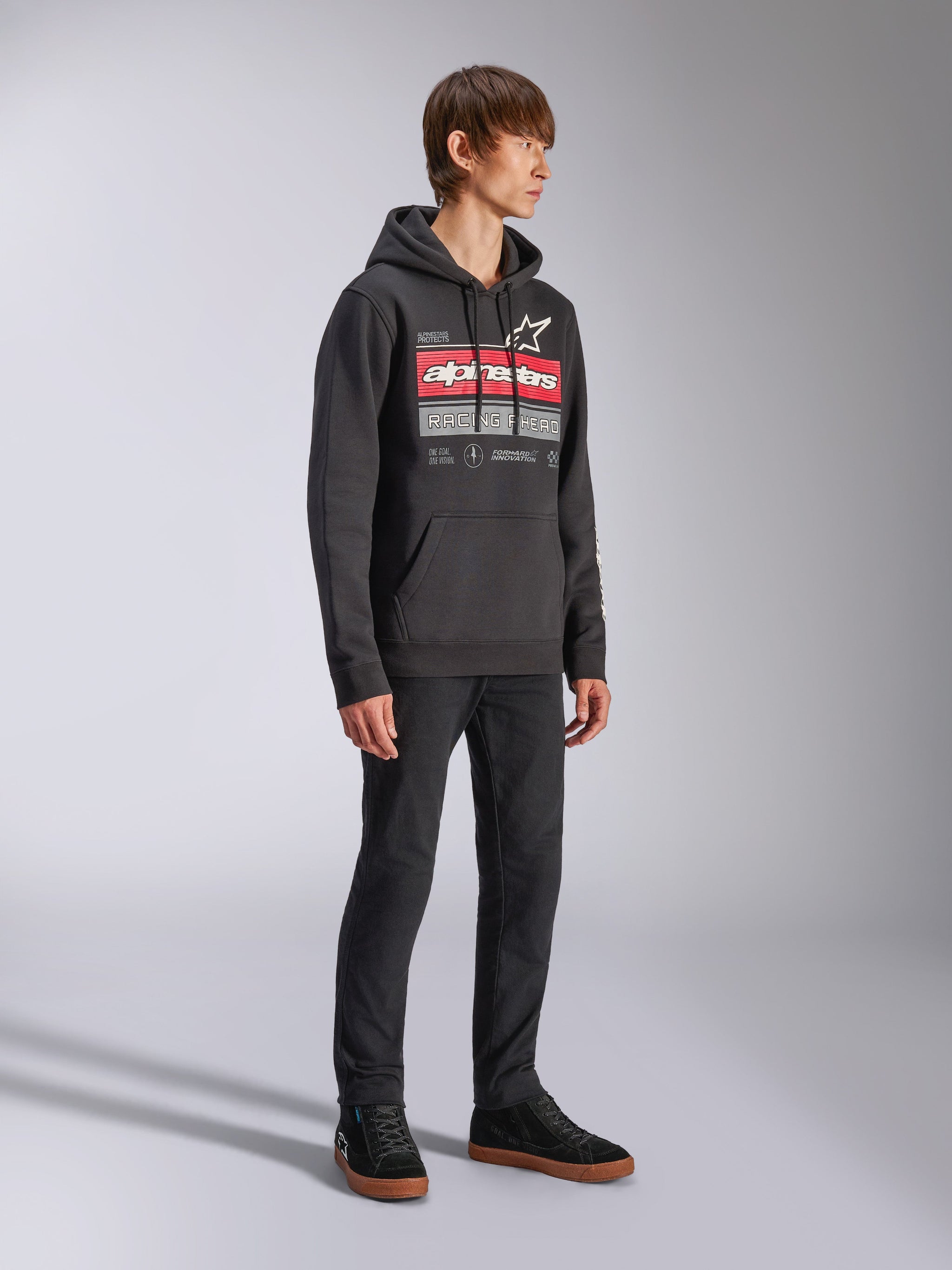 Alpinestars Harken Hoodie, felpa in pile nera con cappuccio, con una grande grafica sul petto con scritta Alpinestars rossa e logo a stella, tasca a marsupio, indossata da un modello con pantaloni neri e sneakers, streetwear casual motorsport