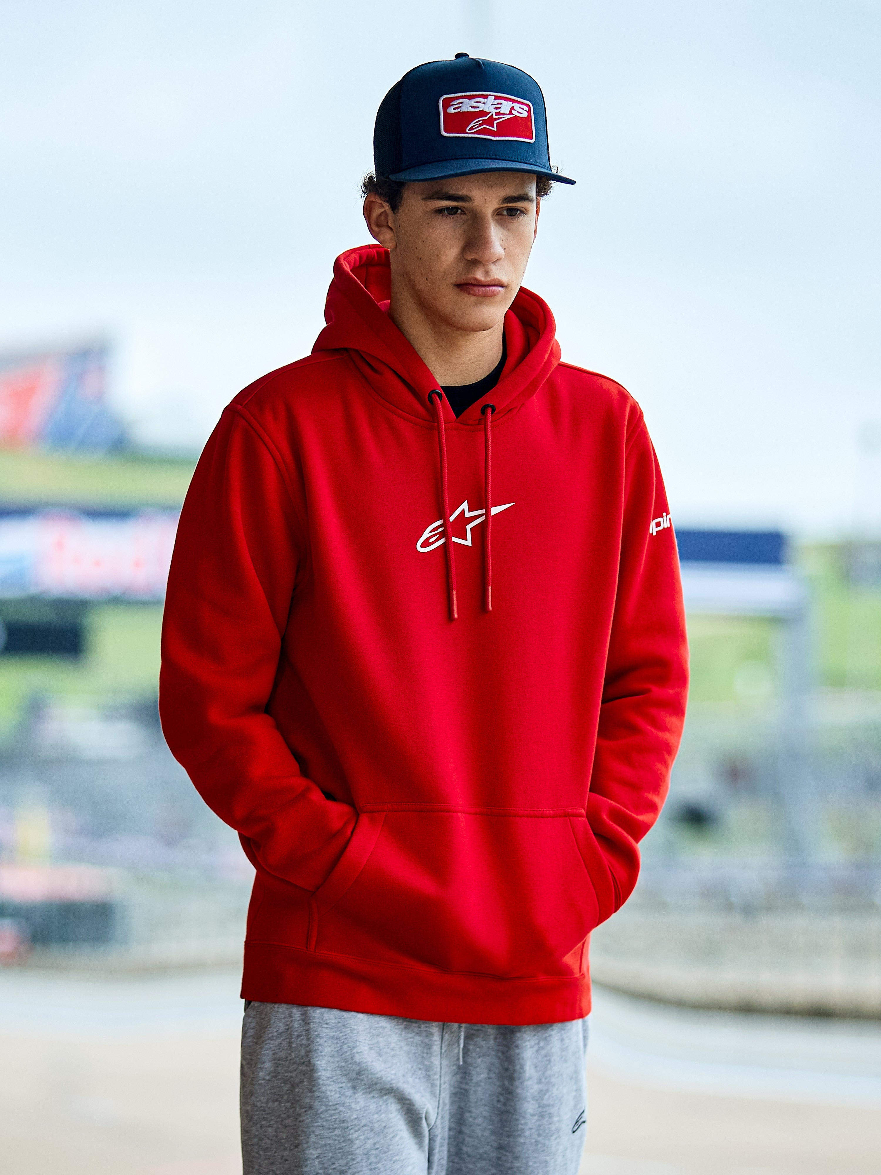 Alpinestars Frontal Hoodie, felpa rossa stile pullover in pile con logo Astars bianco sul petto, tasca a marsupio e cappuccio con cordino, indossata da un uomo con berretto blu, streetwear casual motorsport