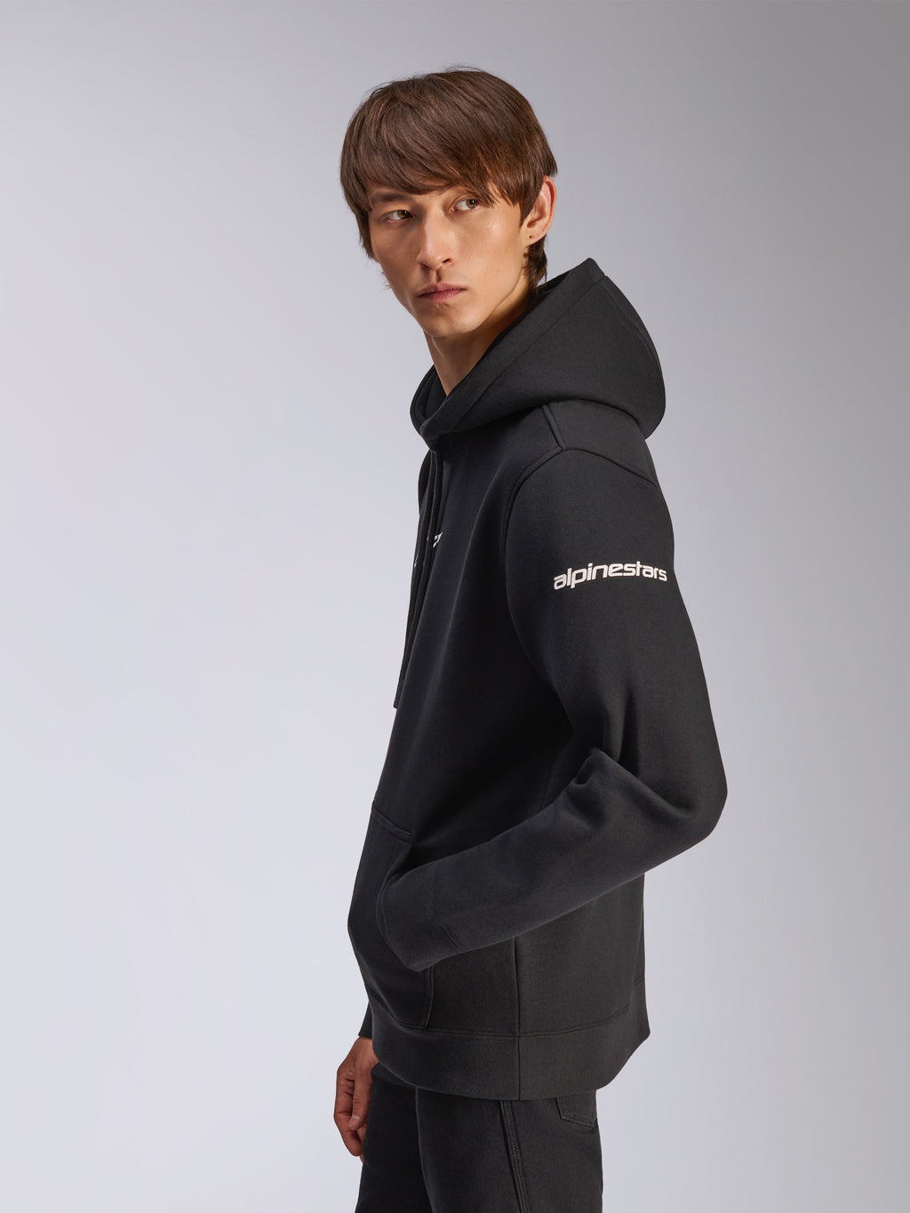 Alpinestars Felpa con cappuccio Frontal, felpa nera in pile, modello ritratto di profilo, logo Alpinestars bianco sulla manica, tasca a marsupio, streetwear casual ispirato al motorsport