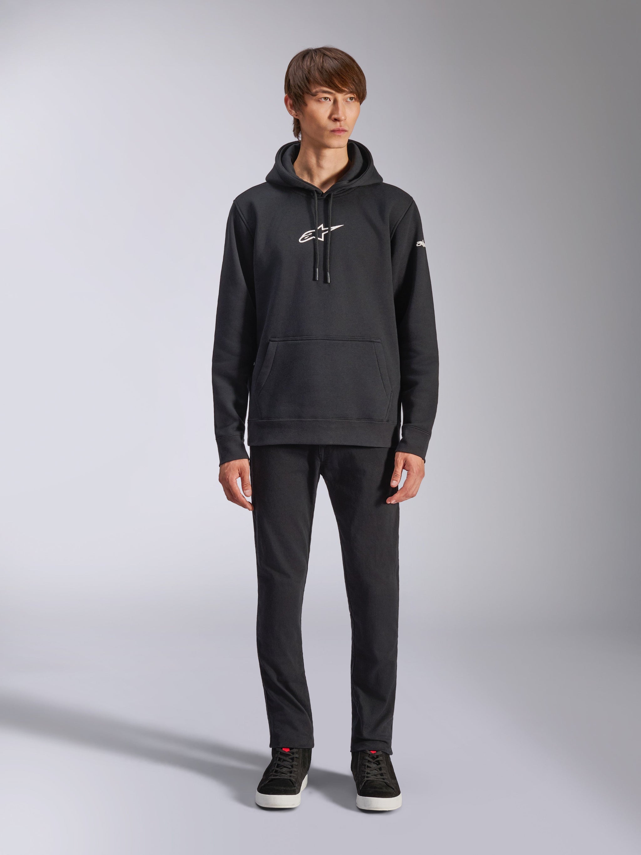 Alpinestars Frontal Hoodie, felpa pullover nera in pile, logo Alpinestars bianco sul petto e sulla manica, tasca a marsupio, cappuccio con cordino, indossata con pantaloni neri, design casual ispirato al motorsport