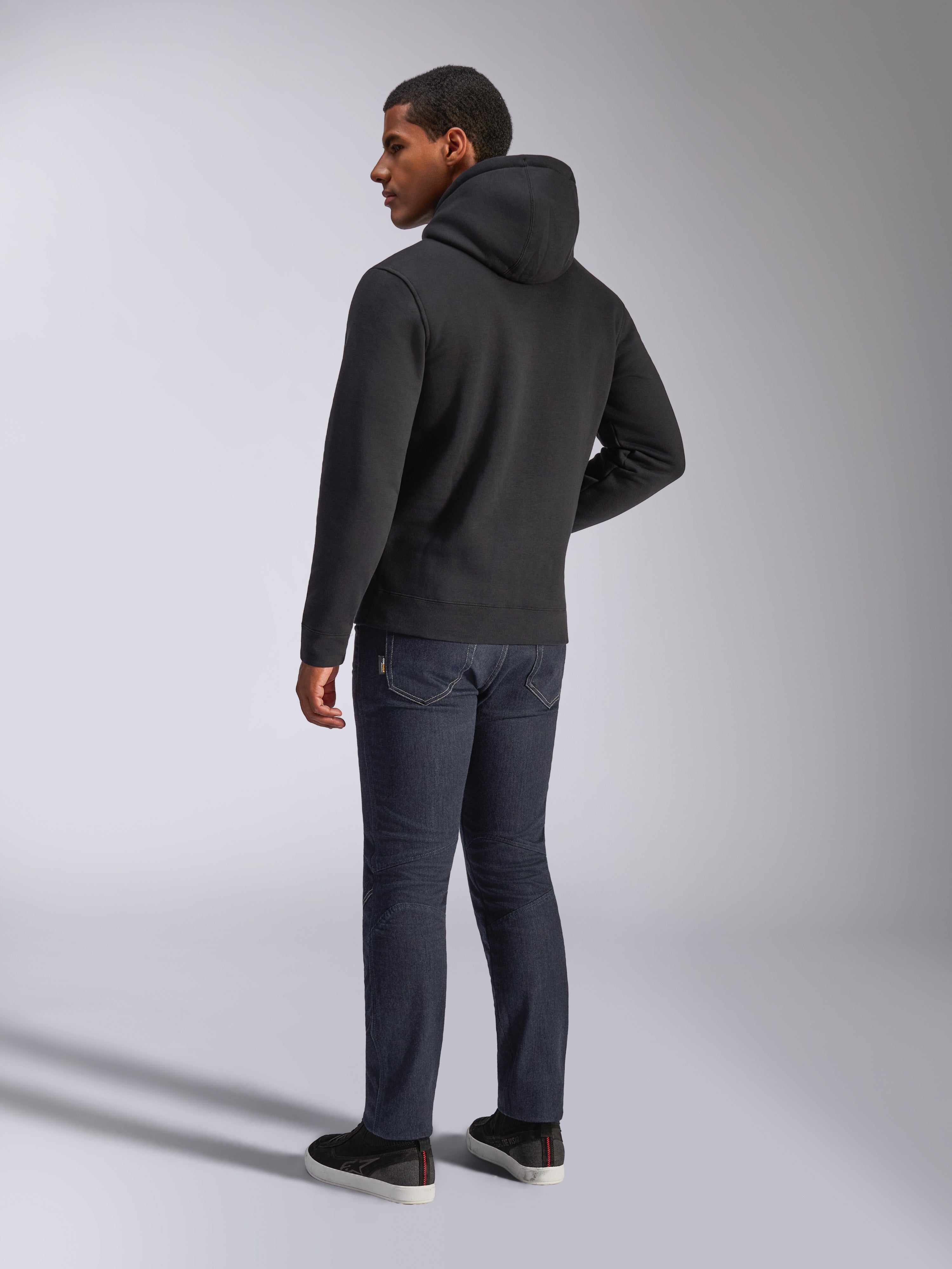 Felpa con cappuccio Alpinestars Blaze V3, vista posteriore di un uomo che indossa la felpa nera, abbinata a jeans scuri e sneakers nere, streetwear casual ispirato al motorsport