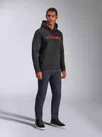 Alpinestars Blaze V3 Hoodie, felpa nera con cappuccio con logo Alpinestars rosso sul petto, indossata da un modello con jeans in denim scuro e sneakers nere, streetwear casual ispirato al motorsport