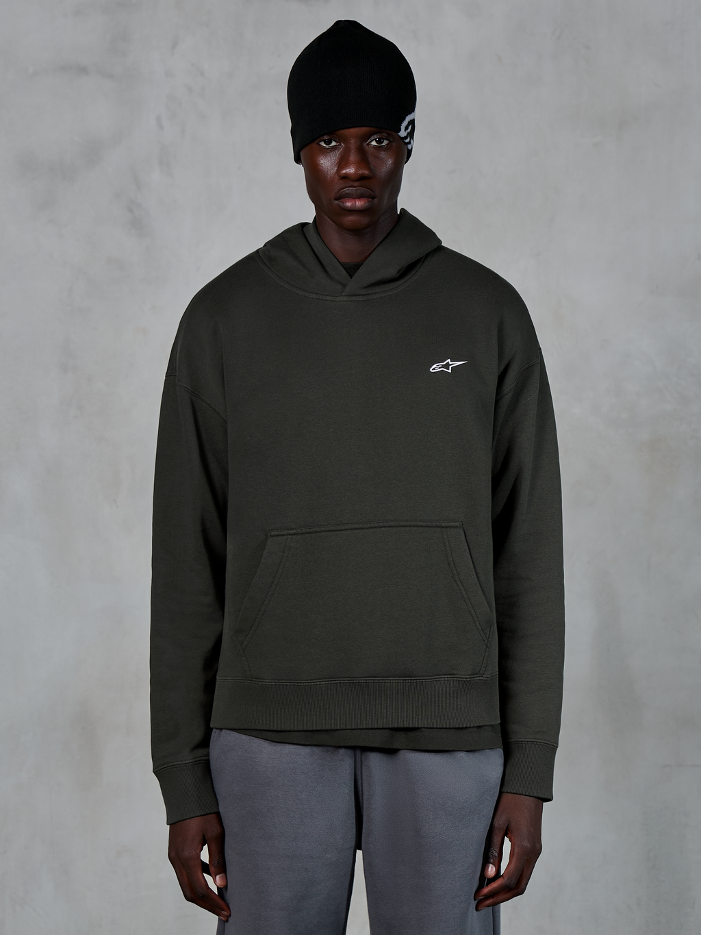 Alpinestars Essentials Ageless Hoodie, streetwear casual, pullover Off Black con un piccolo logo Alpinestars bianco sul petto, indossata con un berretto nero, design minimalista con tasca a marsupio frontale e finitura stone-wash