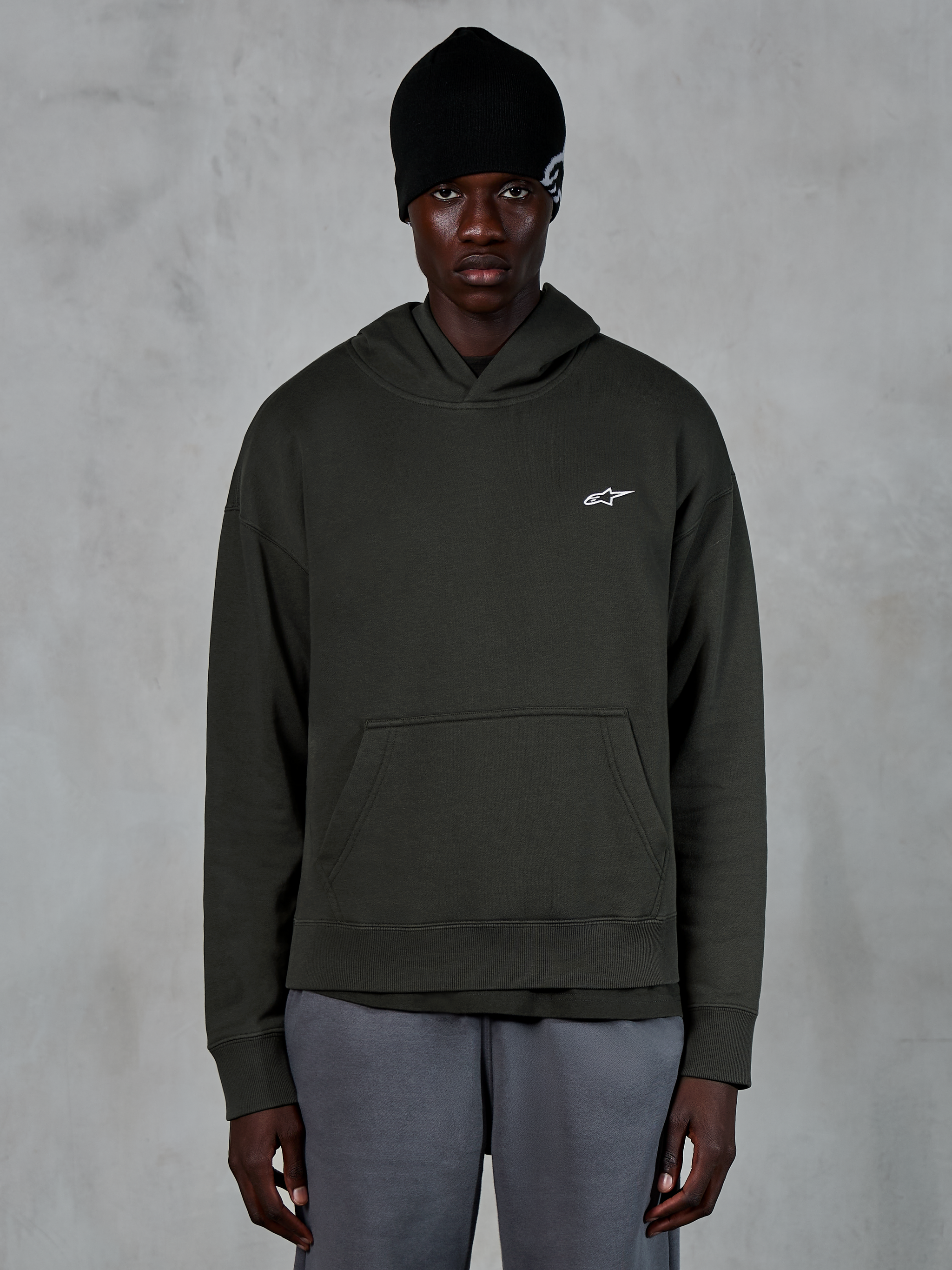 Alpinestars Essentials Ageless Hoodie, streetwear casual, pullover Off Black con un piccolo logo Alpinestars bianco sul petto, indossata con un berretto nero, design minimalista con tasca a marsupio frontale e finitura stone-wash