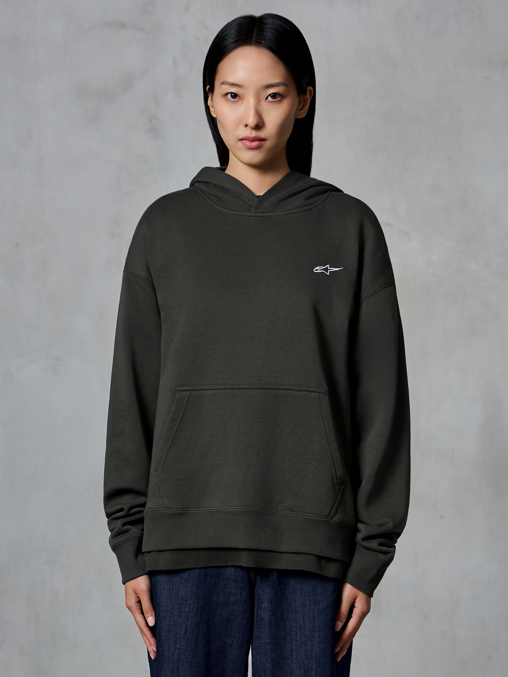 Felpa con cappuccio Alpinestars Essentials Ageless, Off Black, indossata da una modella, con un piccolo logo Astars bianco sul petto, tasca a marsupio frontale, finitura stone-wash, streetwear casual in pile pregiato