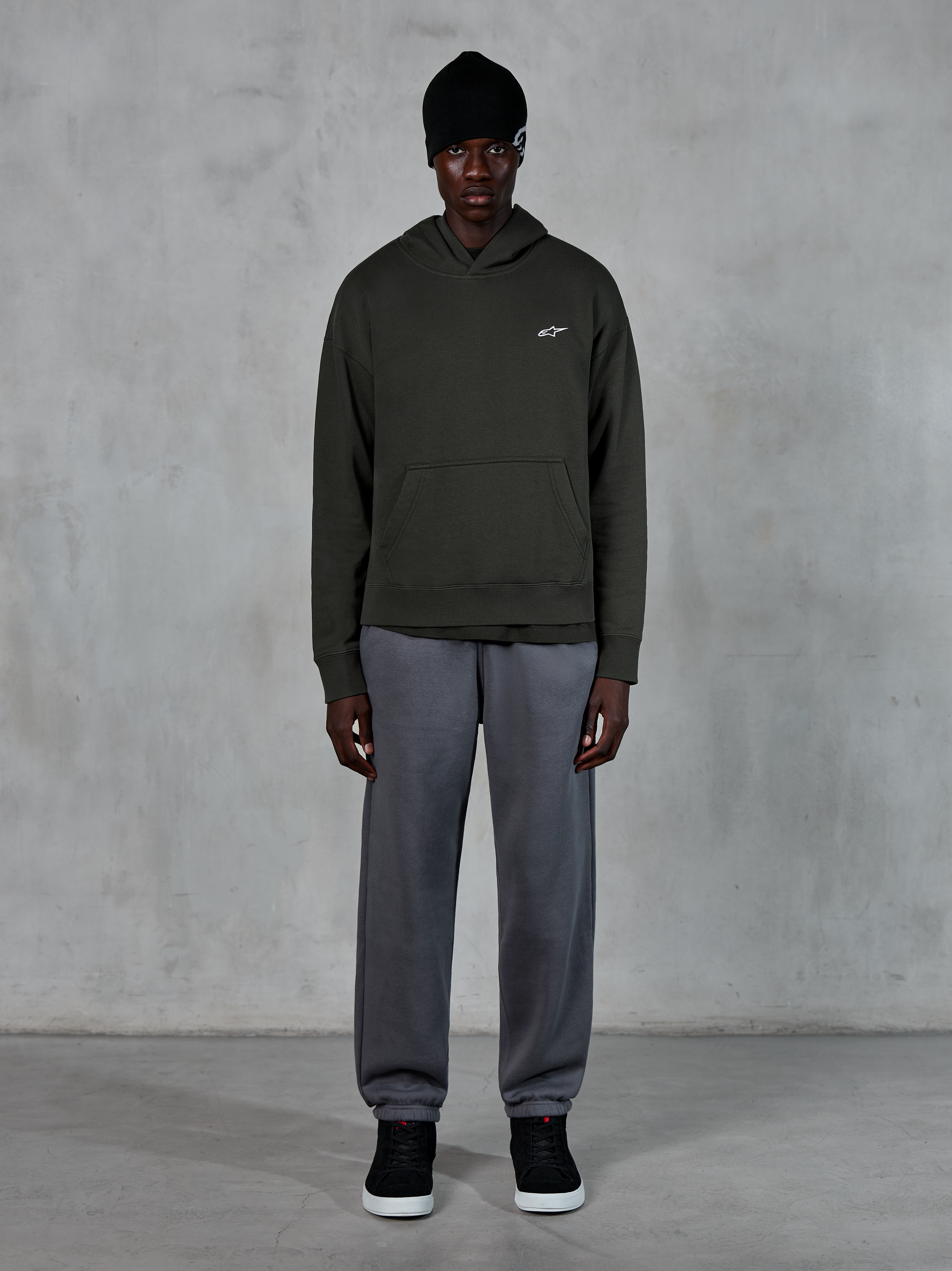 Modello che indossa la felpa con cappuccio Alpinestars Essentials Ageless, streetwear casual, Off Black con piccolo logo bianco sul petto, abbinata a joggers grigi, berretto nero e sneakers nere, design minimalista ispirato al motorsport