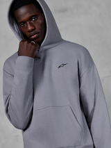 Alpinestars Essentials Ageless Hoodie, argento, modello maschile che indossa la felpa con il cappuccio alzato, piccolo logo Alpinestars nero sul petto, tasca a marsupio, polsini a costine, streetwear casual lifestyle