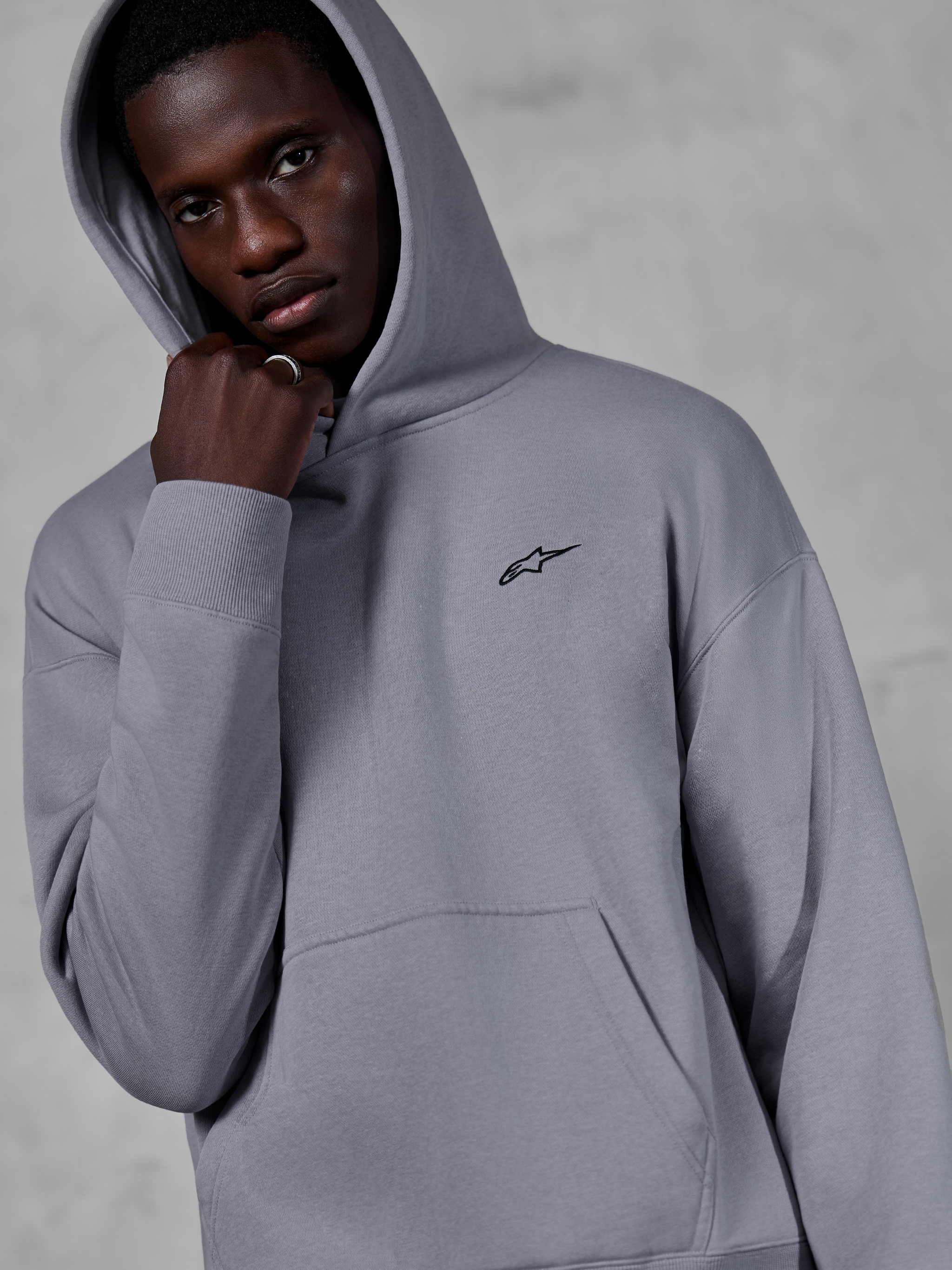 Alpinestars Essentials Ageless Hoodie, argento, modello maschile che indossa la felpa con il cappuccio alzato, piccolo logo Alpinestars nero sul petto, tasca a marsupio, polsini a costine, streetwear casual lifestyle