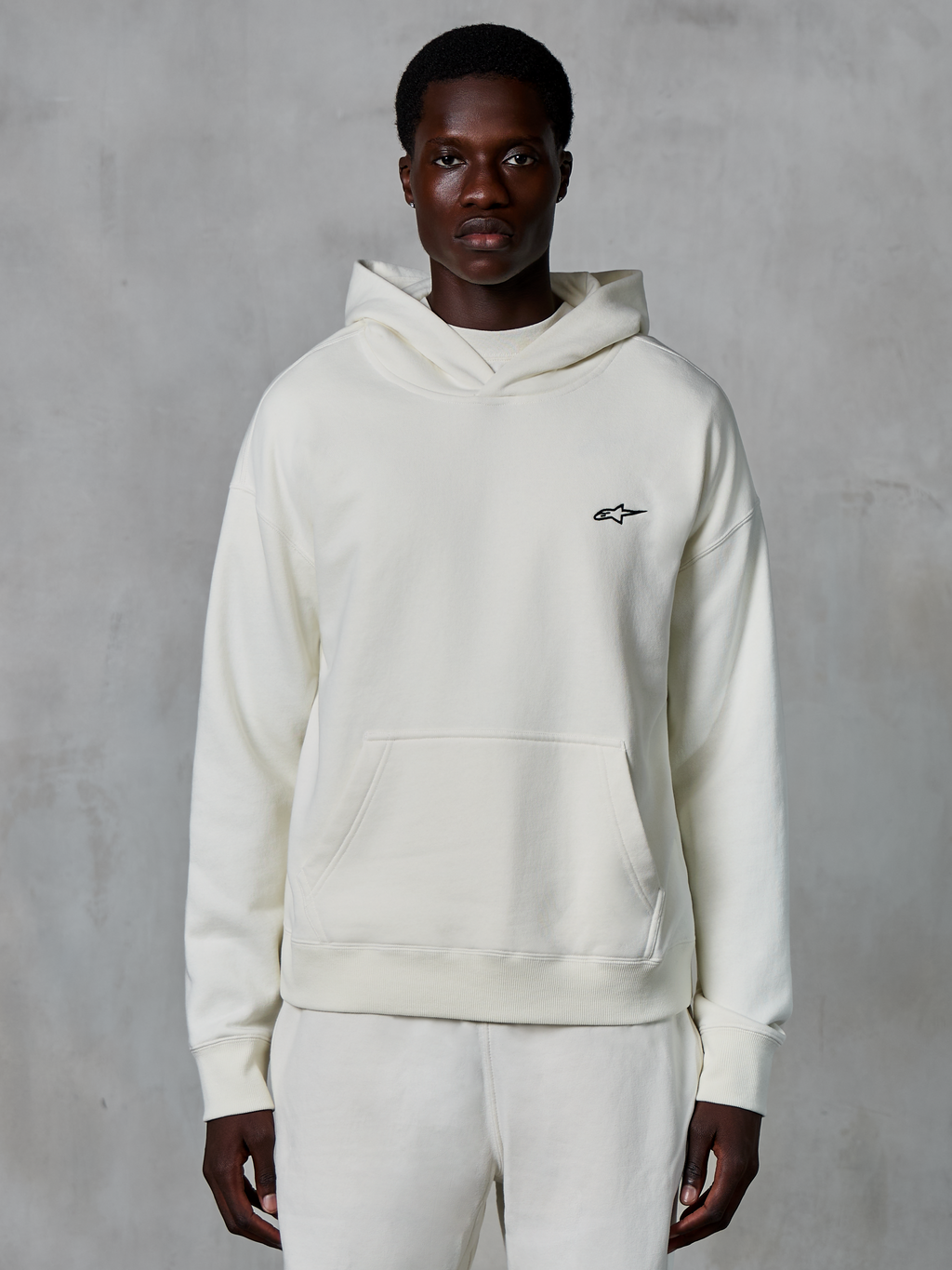 Alpinestars Essentials Ageless Hoodie, bianco sporco, indossata da un modello uomo, piccolo logo nero sul petto, tasca a marsupio frontale, maniche lunghe, streetwear casual ispirato al motorsport