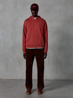 Alpinestars Essentials Ageless Hoodie, felpa casual stile sportswear in colore rosso mattone con finitura stone wash, piccolo logo Alpinestars nero sul petto e tasca a marsupio frontale, indossata da un modello con cappellino bordeaux e pantaloni di velluto a coste marroni per un look streetwear pulito.