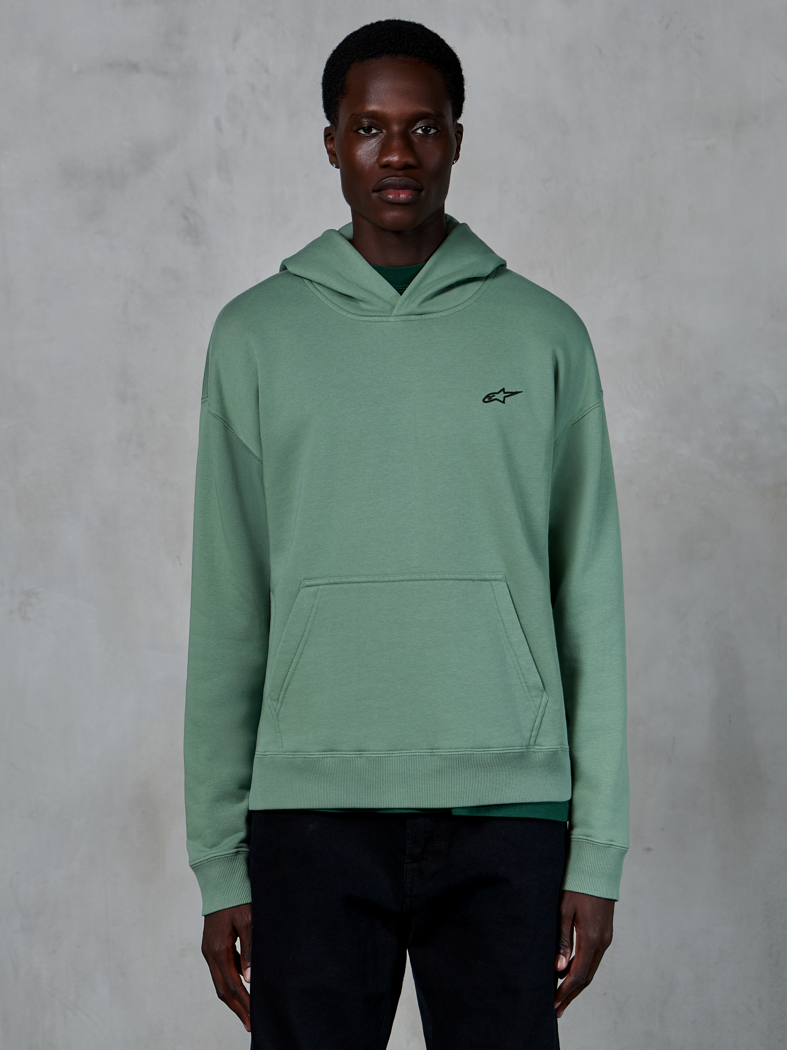 Felpa con cappuccio Alpinestars Essentials Ageless, verde Cactus, felpa casual in pile con finitura stone-wash, con piccolo logo a stella nero sul petto e tasca a marsupio frontale, indossata da un modello in un contesto lifestyle essenziale