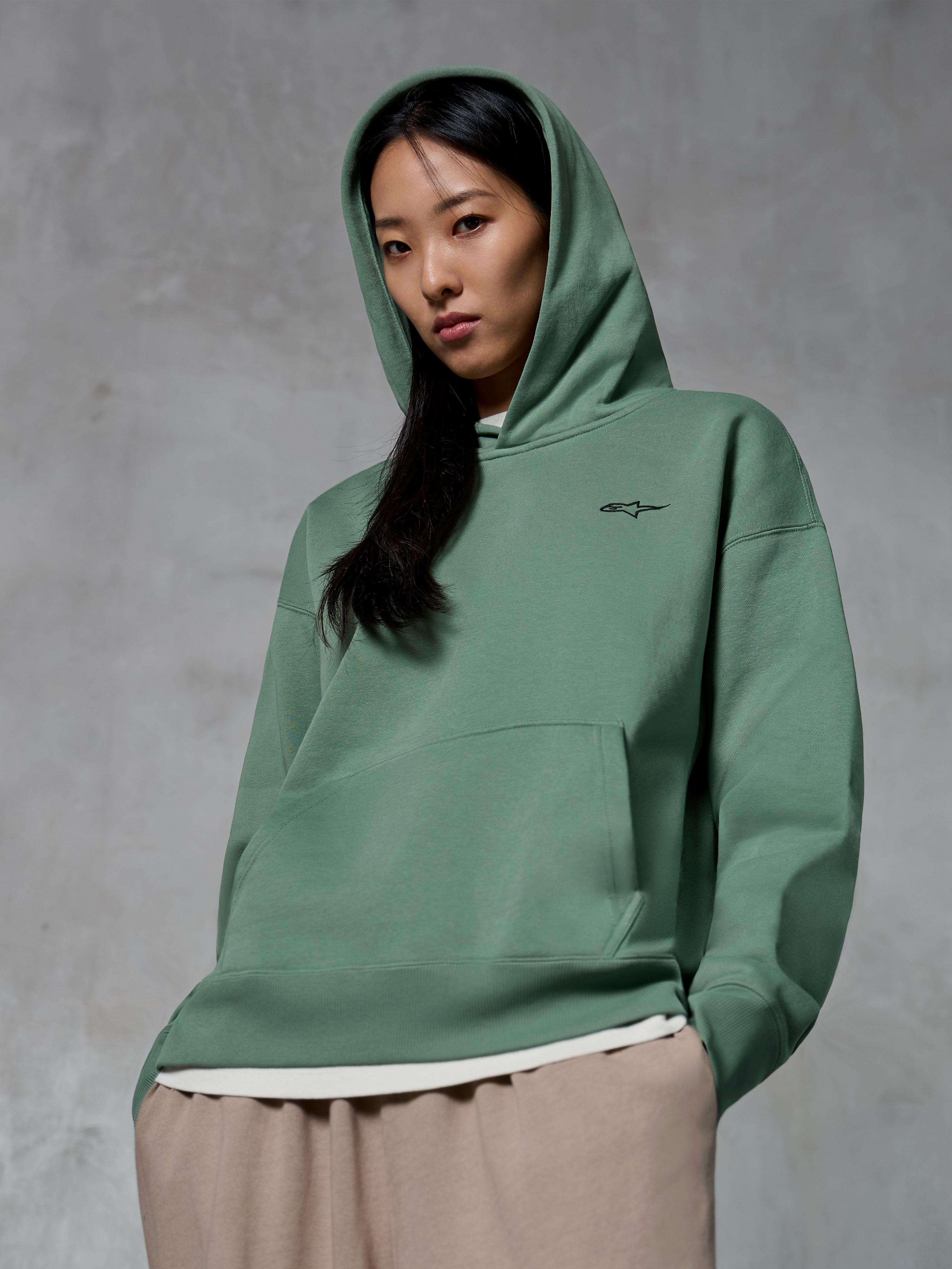 Alpinestars Essentials Ageless Hoodie, streetwear casual, verde Cactus con piccolo logo nero, felpa indossata con cappuccio alzato, tasca a marsupio frontale, finitura stone-wash, abbinata a pantaloni beige chiaro